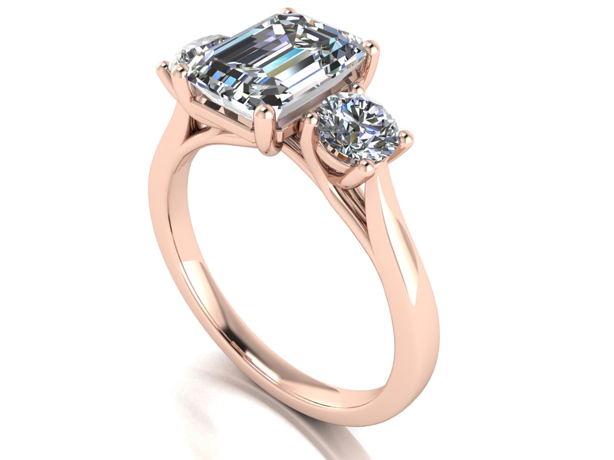 angled view of a trilogy rose gold engagement ring set with an emerald centre lab grown diamond with two round brilliant diamonds on either side of the centre stone